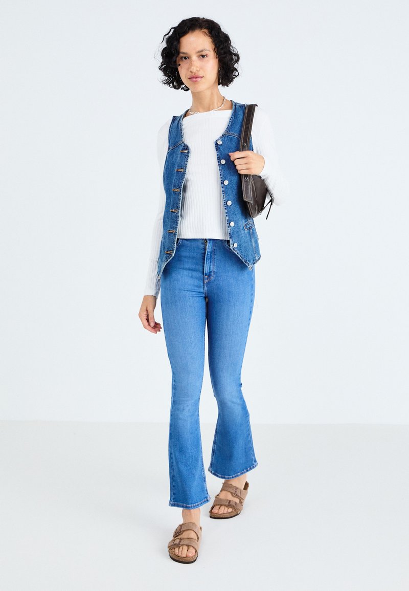 Jeansweste mit Knopfleiste, kombiniert mit einem gerippten Langarmoberteil und blauen Schlagjeans. Getragen mit braunen Sandalen und einer schwarzen Schultertasche.