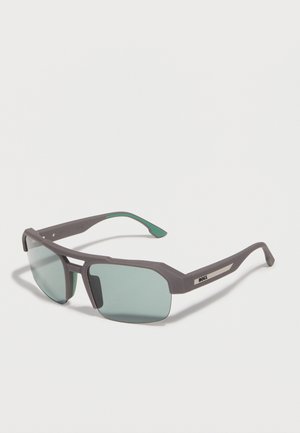 Gafas de sol rectangulares con montura gris, lentes de tono verde y un detalle metálico en las patillas, colocadas sobre una superficie blanca en ángulo.