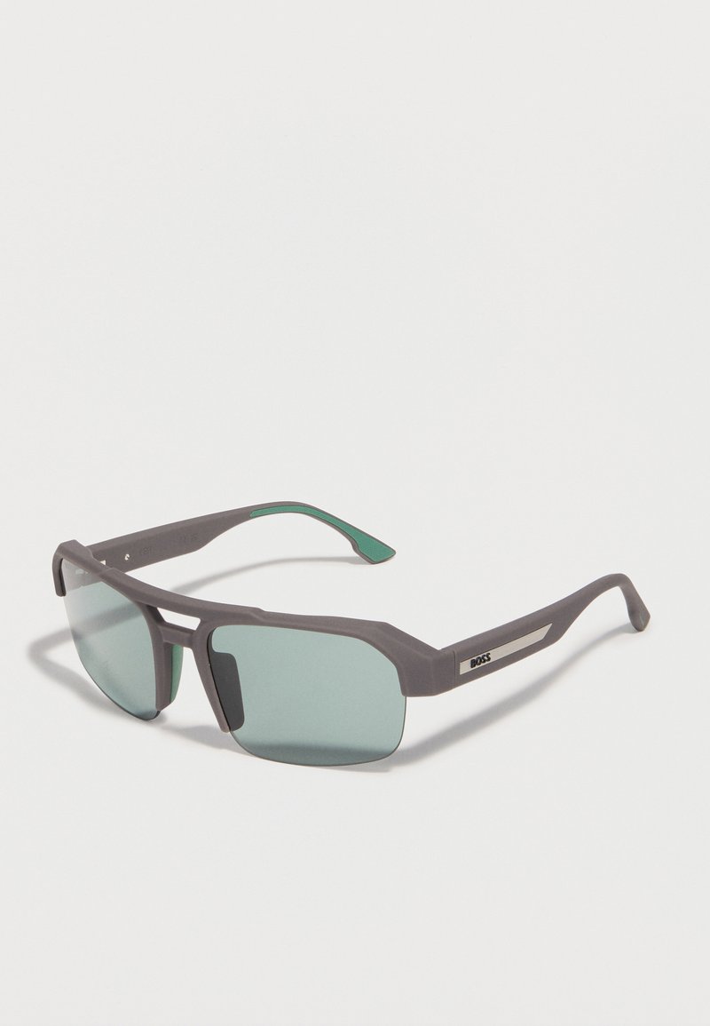 Lunettes de soleil à monture rectangulaire grise avec verres teintés verts et accent métallique sur les branches, posées sur une surface blanche en angle.
