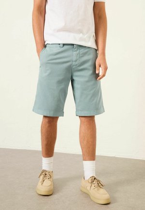 Man wearing light blue knee-length shorts, white socks, beige lace-up shoes, and a white t-shirt, standing indoors on a gray floor.