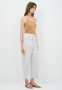 Een tan spaghetti-top gecombineerd met loszittende lichtgrijze broek met zijzakken, gestyled met platte sandalen. De achtergrond is helemaal wit.