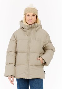 Vrouw in een beige pufferjack met ritszakken en capuchon, gecombineerd met een beige gebreide beanie en blauwe jeans, recht vooruit kijkend.
