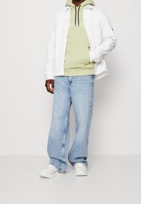 Vit denimjacka över en ljusgrön hoodie med svarta snören, tillsammans med ljusblå vida jeans och vita sneakers.