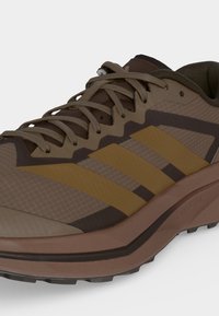 Καφέ και ανοιχτό καφέ αθλητικό παπούτσι με κορδόνια, τρεις κίτρινες πλευρικές ρίγες και σχέδιο σόλας για υπαίθρια χρήση.