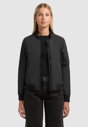 Mladá žena v čiernej bombera bundě přes čierne tričko a čierne rifle, stojaca pred jednoduchým svetlým pozadím.