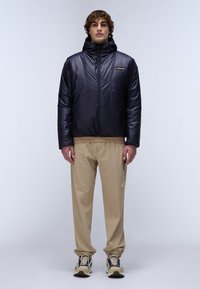 Marineblauwe pufferjack met een hoge kraag en een ritssluiting aan de voorkant, gecombineerd met beige broek met elastische taille en beige en zwarte sneakers.