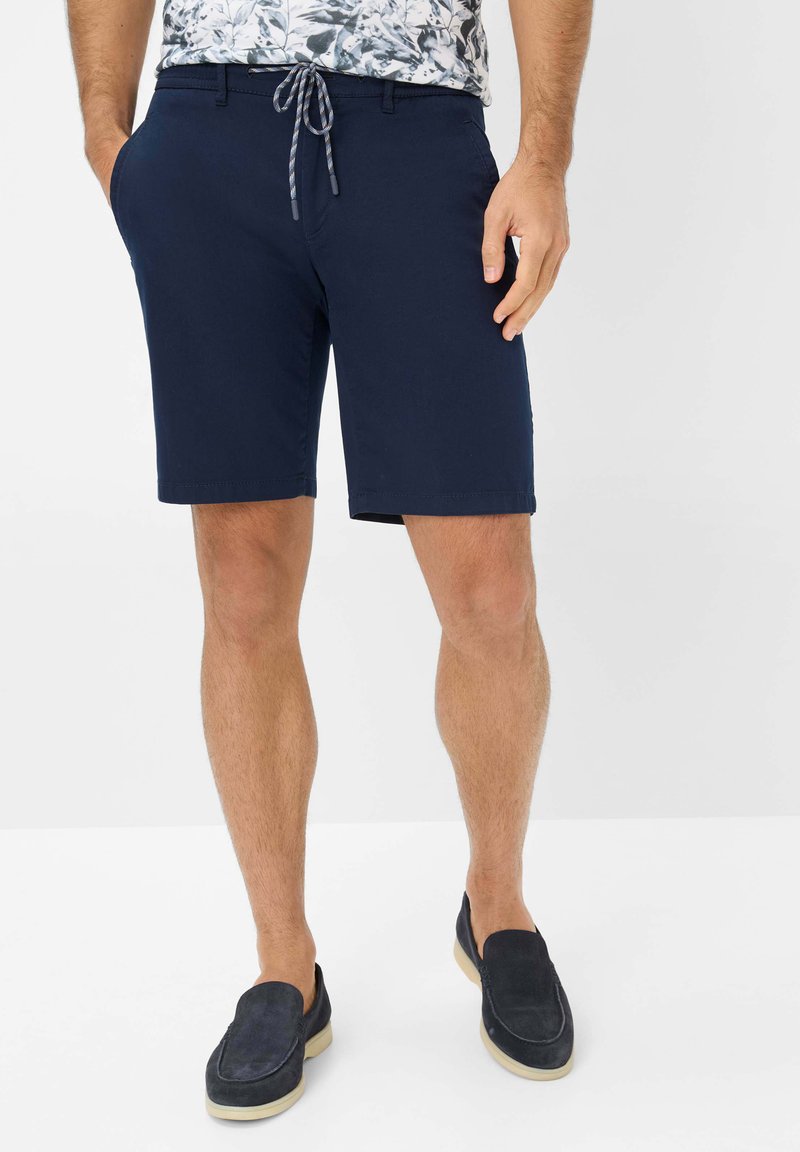 Bermuda blu navy con elastico in vita, tasche laterali e una vestibilità comoda, abbinate a scarpe slip-on scure e una camicia a fantasia floreale.