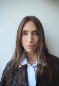 Chaqueta de cuero marrón con una camisa blanca de cuello por debajo, con cabello largo y liso marrón y mirada directa frente a un fondo claro.