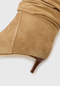 Stivaletto in suede beige con design morbido, tacco affusolato e texture liscia. Presenta una cucitura lungo il lato per un tocco di dettaglio in più.