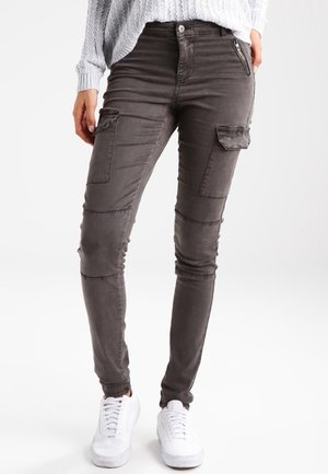 Pantalon cargo gris, coupe slim, avec poches zippées et finition lisse légèrement texturée. Associé à des baskets blanches.