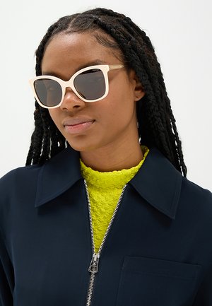 Donna con lunghi capelli intrecciati che indossa occhiali da sole oversize rosa chiaro, un top verde lime testurizzato e una giacca blu navy con zip.