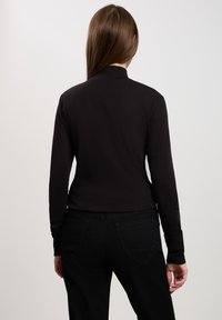 Czarny top z długim rękawem z kołnierzykiem stójkowym, gładką fakturą i dopasowanym krojem, w połączeniu z czarnymi dżinsami z tylnymi kieszeniami i subtelnym przeszyciem.