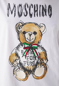 Ilustracja misia pluszowego w białej koszulce z napisem "EURE FOR FUN" oraz czerwoną, zieloną i białą kokardą, z napisem "MOSCHINO" powyżej.