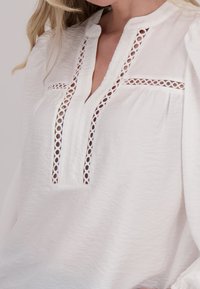 Witte blouse met een kraagloos ontwerp, ingewikkelde oogjesaccenten en een gekreukte textuur. Lange mouwen met gathered manchetten.