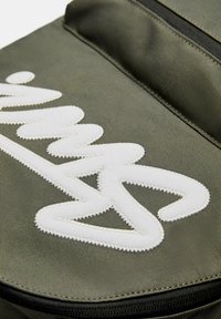 Olivgrön tygryggsäck med en framträdande vit broderad logotyp. Har en slät yta och dragkedjestängning i toppen.