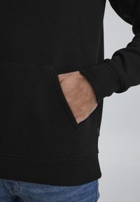 Felpa nera in misto cotone, dotata di tasca a marsupio, polsini e orlo ribbed. Etichetta visibile sul lato. Dettaglio ravvicinato.