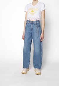 Avklippt vit T-shirt med gult Adidas-logotyp, tillsammans med vida blå jeans och beige sneakers. Jeansen har hög midja och synliga sömmar.