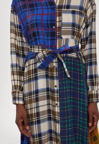 Robe boutonnée à manches longues avec des motifs à carreaux mélangés en bleu, vert, marron et crème, nouée à la taille avec une ceinture en tissu assorti.