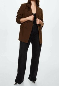 Femme portant un blazer marron à double boutonnage, un haut noir, un jean noir taille haute à jambes larges et des chaussures argentées pointues.