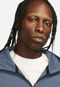 Blue zip-up jacket with black accents, layered over a white shirt. Braided hair with metallic accessories and facial piercings visible.
