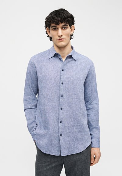 Νεαρός άνδρας με σγουρά σκούρα μαλλιά, φορώντας ανοιχτό μπλε πουκάμισο με κουμπιά και σκούρο γκρι παντελόνι, στέκεται μπροστά σε απλό λευκό φόντο.