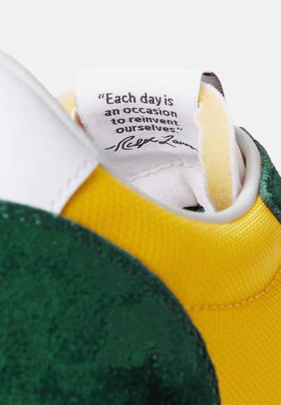 Πολύχρωμο αθλητικό παπούτσι με πράσινο σουέντ, κίτρινο ύφασμα και λευκές λεπτομέρειες. Η ετικέτα στο εσωτερικό εμφανίζει ένα παρακινητικό απόφθεγμα με μαύρο κείμενο.