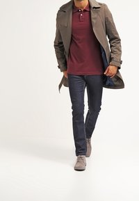 Manteau trench en brun par-dessus un polo bordeaux, jean foncé et chaussures à lacets grises. Tissu lisse, coupe ajustée et style décontracté.