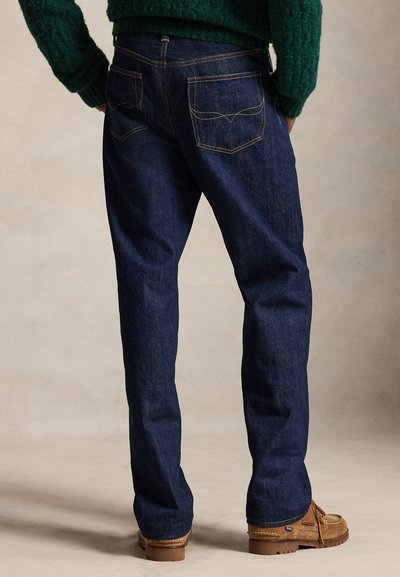 Persona con vaqueros rectos azul oscuro, suéter de punto verde y zapatos mocasines marrones, de pie sobre un fondo neutro.