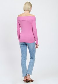 Rosa, schulterfreier Pullover mit langen Ärmeln, geripptem Saum und glatter Textur, kombiniert mit hellblauen Jeans und braunen, hohen Pantoletten.