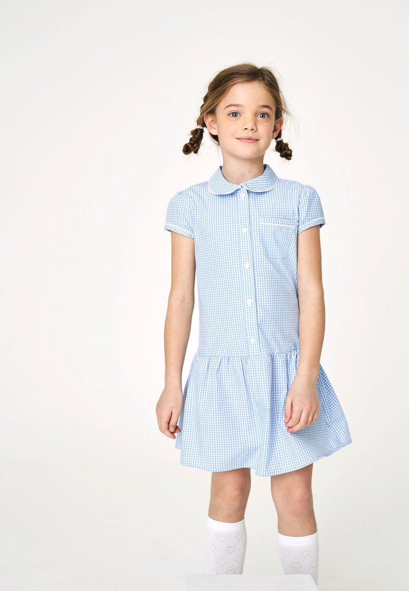 Vestido de cuadros azul con cuello tipo camisero, mangas cortas abullonadas, botonera frontal y falda con volantes. Combinado con calcetas blancas hasta la rodilla con un patrón de corazones.