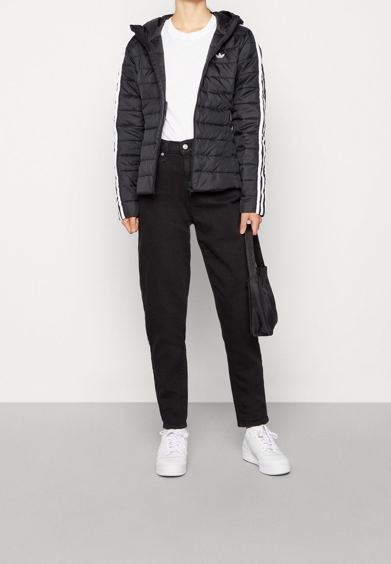 Zwarte pufferjack met een capuchon en witte streepdetails, gecombineerd met zwarte broek en witte sneakers. Het model houdt een zwarte handtas vast.