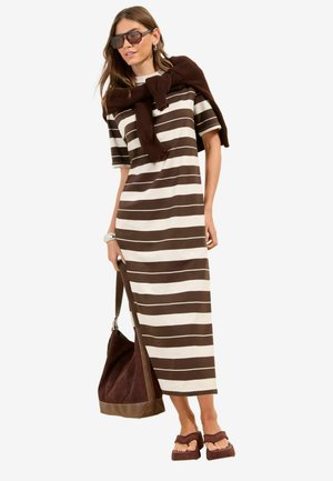 Naine, kes kannab pruuni ja valge triibulist midi kleiti, tumedad päikeseprillid, kampsun üle õlgade visatud, kaasas pruun käekott ja pruunid sandaalid.