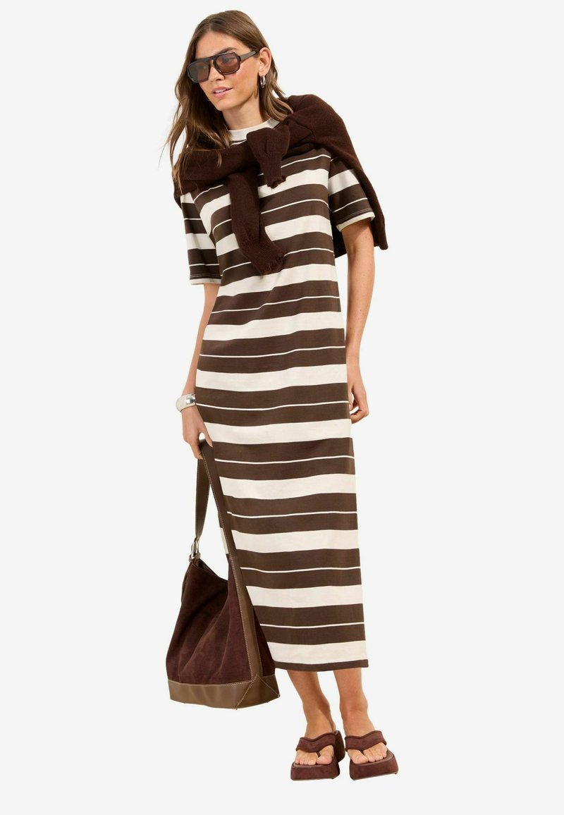 Vrouw die een midi-jurk met bruin-witte strepen draagt, donkere zonnebril, trui over de schouders, draagt een bruine shopper en bruine sandalen.