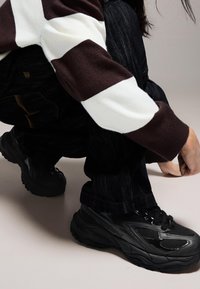 Svarta sneakers med mesh- och gummidetaljer, med en tjock sula, kombinerade med mörka jeans och en randig tröja i brunt och vitt.