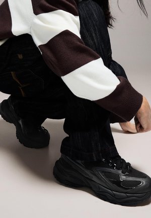 Fekete sportcipők hálós és gumis díszítéssel, vastag talppal, sötét farmerrel és barna-fehér csíkos pulóverrel párosítva.