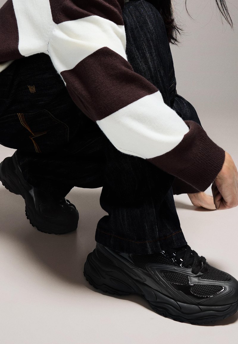 Svarta sneakers med mesh- och gummidetaljer, med en tjock sula, kombinerade med mörka jeans och en randig tröja i brunt och vitt.