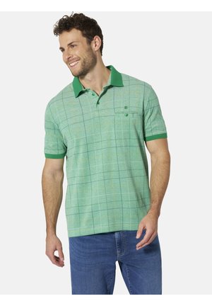 Mann lächelt, trägt ein grün kariertes Poloshirt mit dunklerem grünem Kragen und Ärmelsäumen und blaue Jeans, vor einfarbigem Hintergrund.