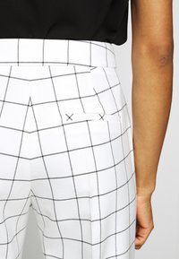 Pantalon blanc à motif quadrillé noir, avec une taille haute et un détail discret de poche. Le tissu présente une texture lisse et structurée.