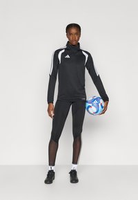 Μαύρο αθλητικό τοπ με λευκές λεπτομέρειες και ημισφαιρικό φερμουάρ, συνδυασμένο με μαύρο κολάν με ρίγες στο πλάι. Κρατώντας μια μπλε και λευκή ποδοσφαιρική μπάλα.