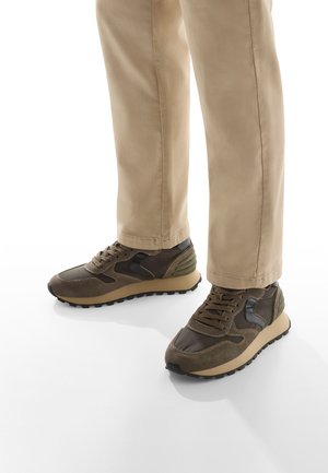 Sneaker marroni con un mix di suede e materiali sintetici, punta rotonda, suola testurizzata e accenti a contrasto sui lati. Pantaloni beige visibili.
