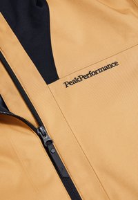 Giacca color marrone chiaro con dettagli neri, caratterizzata da una chiusura con zip e dal logo "PeakPerformance". Tessuto liscio e linee pulite nel design.
