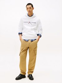 White hoodie with "TOMMY HILFIGER" logo, beige cargo pants with side pockets, and black shoes. Model stands against a neutral background.