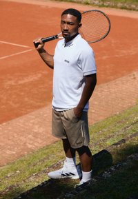 White polo shirt with black "BOSS" logo, beige shorts with pocket detail, white sneakers, holding a tennis racket, standing on red court.
