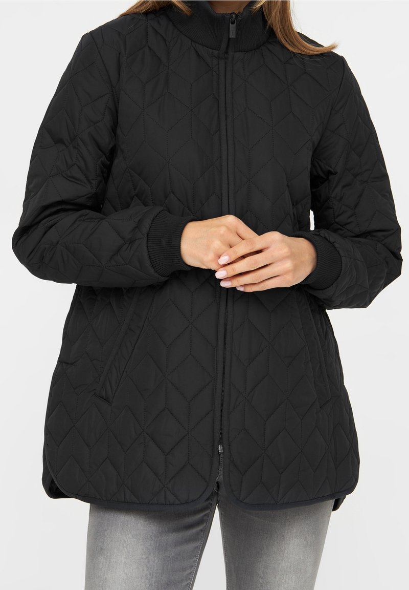 Kvinna klädd i en svart vadderad jacka med diamantstygning, dragkedja, ribbade ärmslut och framfickor, som är kombinerad med urtvättade grå jeans.