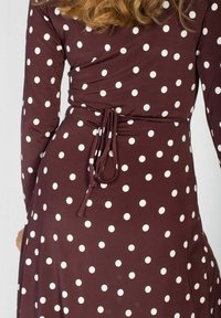 Brązowa sukienka w białe kropki, z długimi rękawami i związanym paskiem w talii, pokazana od tyłu na stojącej osobie.