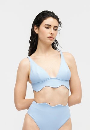 Femeie cu păr întunecat ud, purtând un costum de baie bleu cu talie înaltă și margini ondulate, pe un fundal alb simplu.