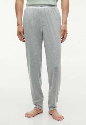 Mężczyzna w jasnoszarych spodniach dresowych Tommy Hilfiger z logo na elastycznym pasie, stojący boso na białym tle.