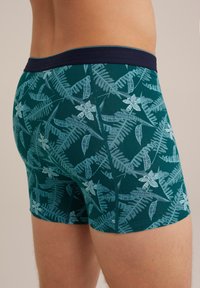 Herrboxershorts i mörkgrön med ett blommigt ormbunksmönster. Slättyg, elastisk midja i marinblått, figurnära design, längd till mitten av låret.