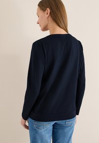 Haut bleu marine à manches longues avec un col rond, texture lisse, coupe droite et détails de couture subtils, associé à un jean bleu clair.