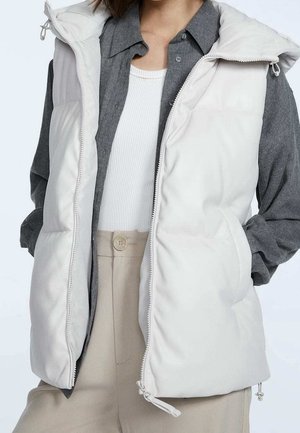 Femme portant une veste sans manches matelassée blanche sur une chemise grise boutonnée et un débardeur blanc côtelé, associée à un pantalon beige taille haute.